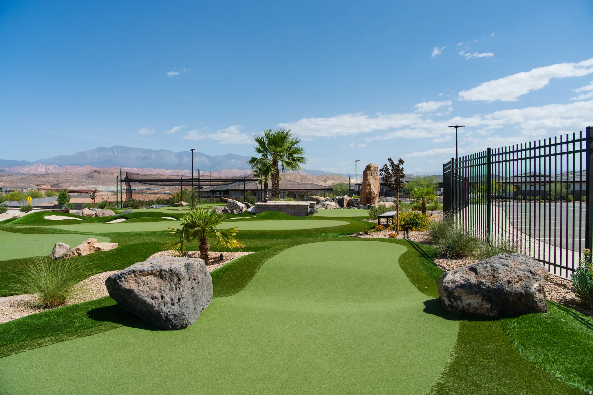 Expansive putting green featuring meticulously manicured synthetic turf, strategically placed boulder accents, mature palm trees, and black metal perimeter fencing