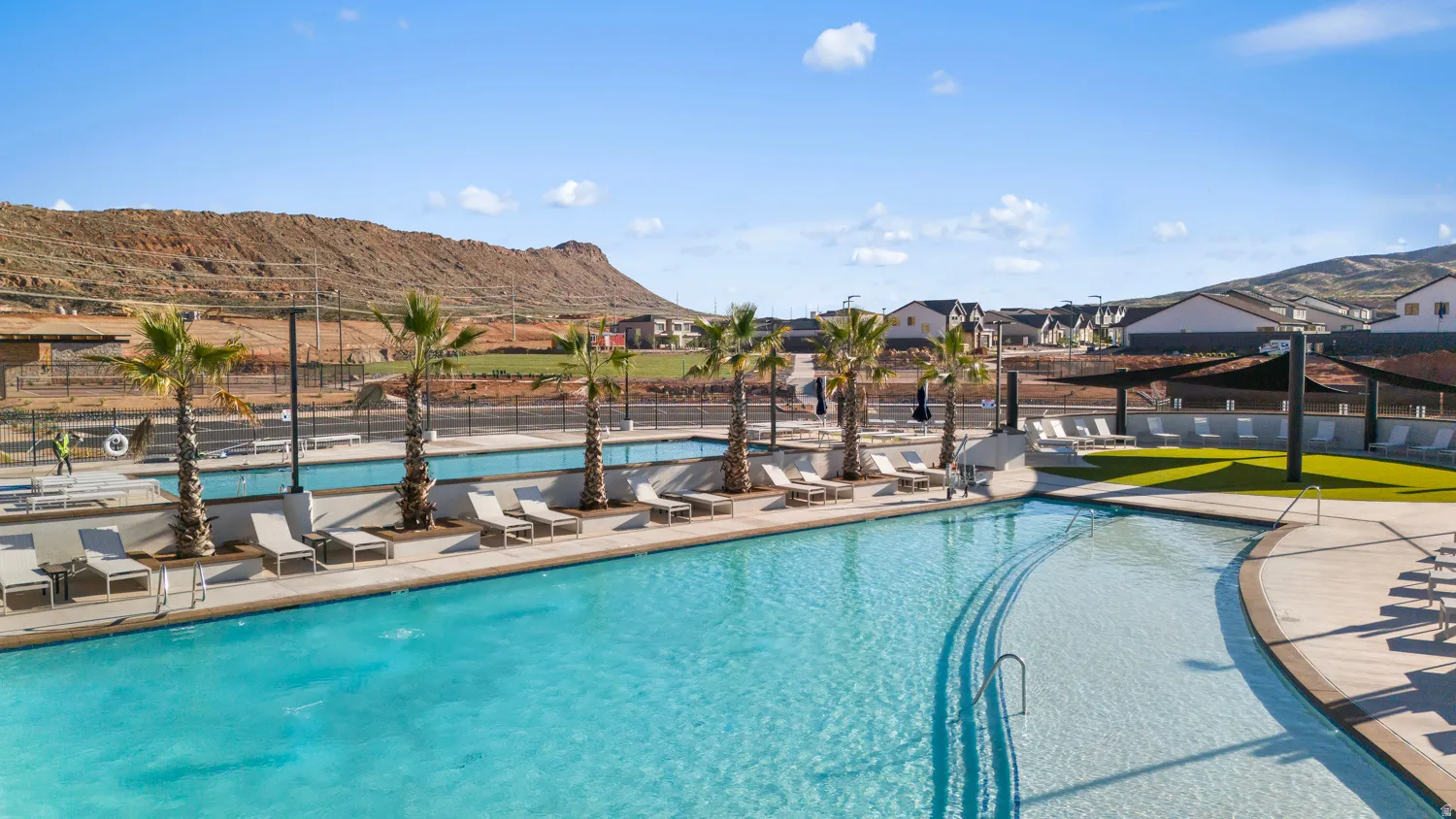 Community pool area featuring a curved swimming pool, multiple palm trees, an adjacent turf lounge area, and a shaded outdoor seating zone