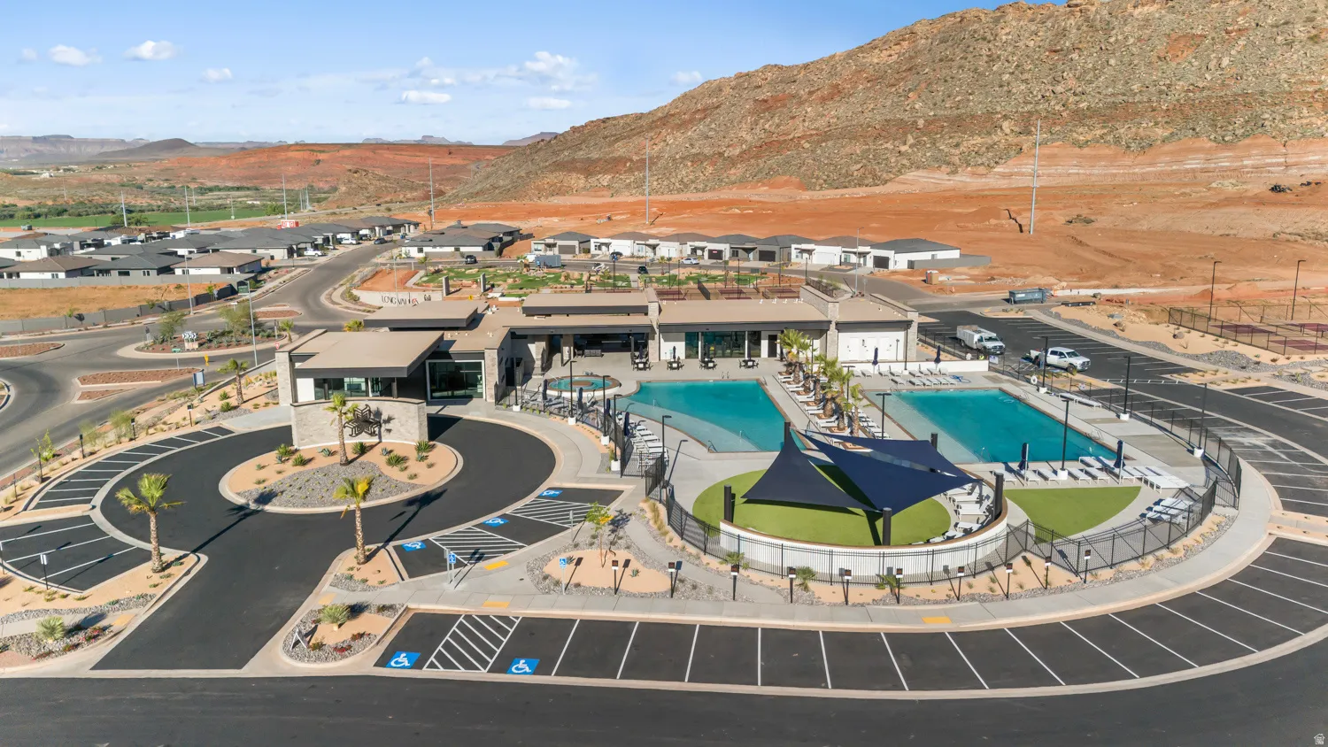 Aerial View of Resort-Style Community Amenities featuring a Resort-Style Swimming Pool with Turquoise Waters, Integrated Spa/Hot Tub with Modern Shade Structure, Professionally Designed Hardscaping and Circular Driveway, Expansive Parking Areas, and Surro