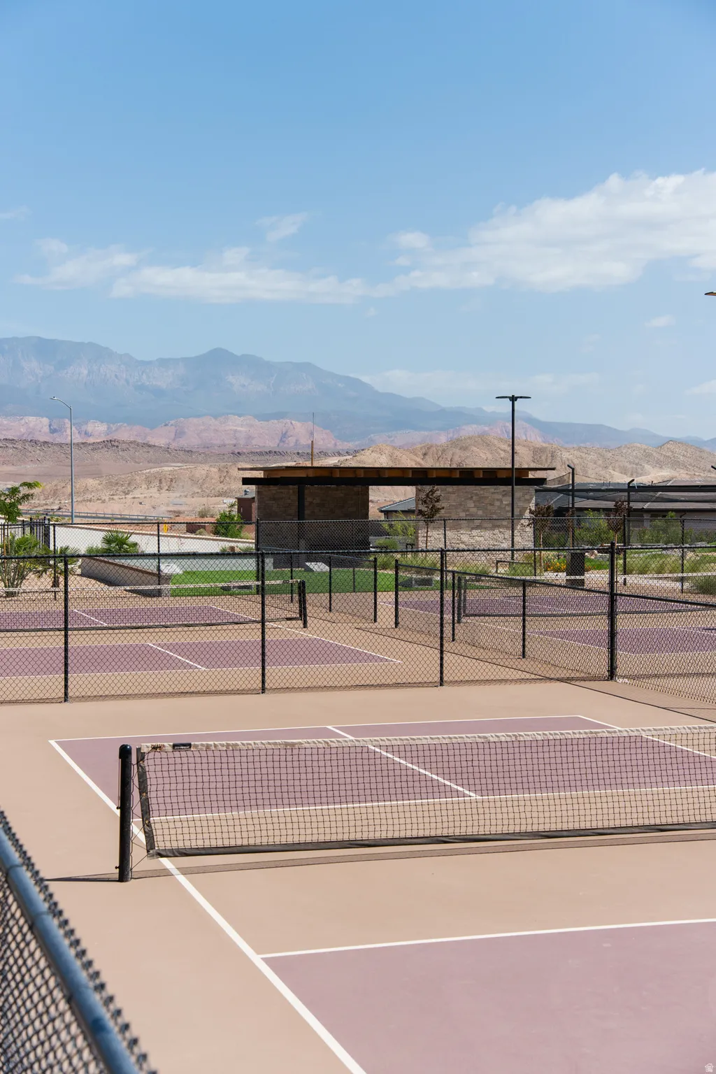 Professional tennis court complex featuring multiple well-lit courts with regulation net systems, durable clay-composite playing surfaces in contrasting earth tones, sturdy perimeter fencing, and a covered pavilion structure with electrical infrastructure