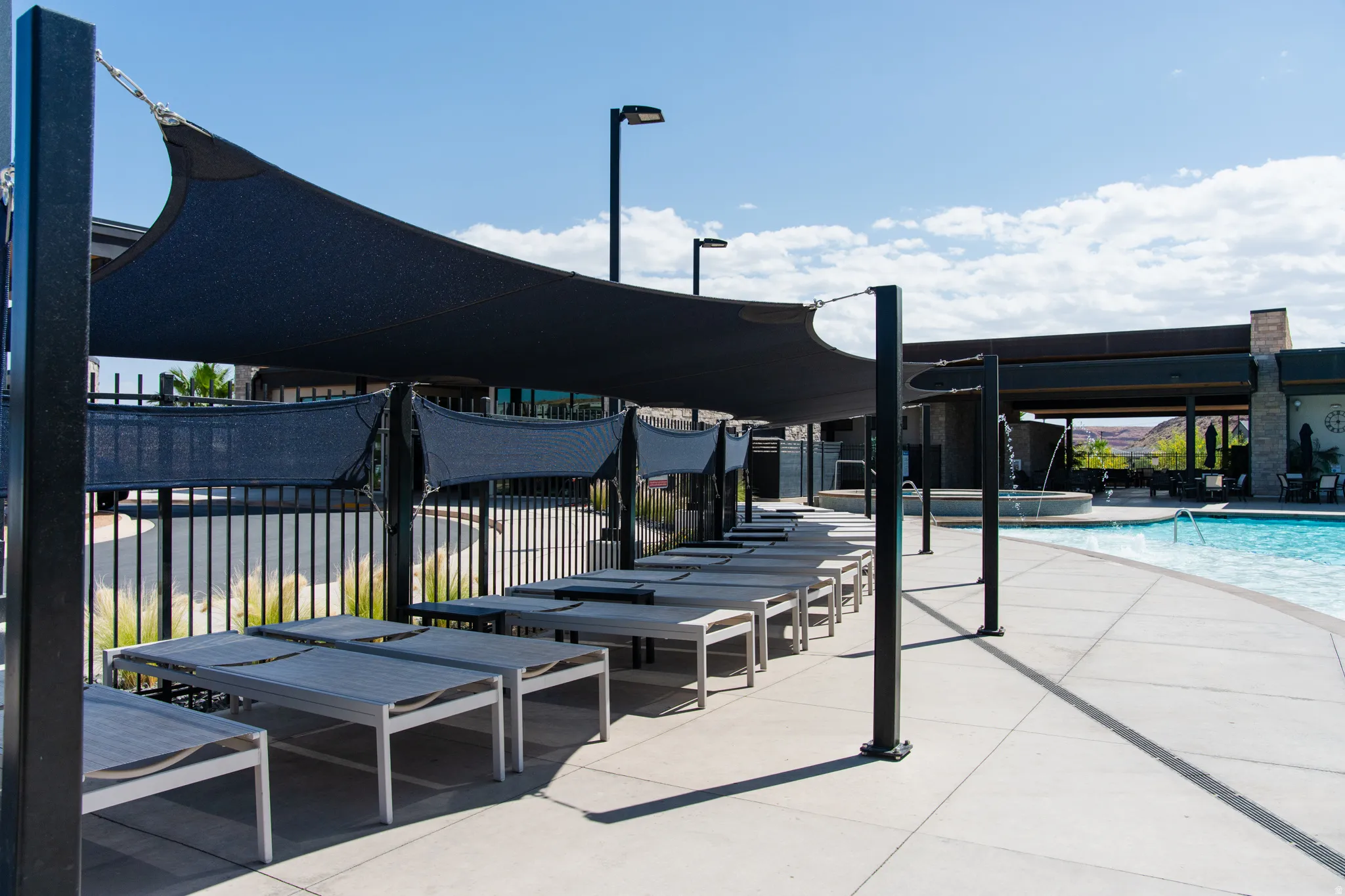 Poolside lounge area featuring a shade sail structure, concrete decking, an adjacent swimming pool, and a stone-clad building