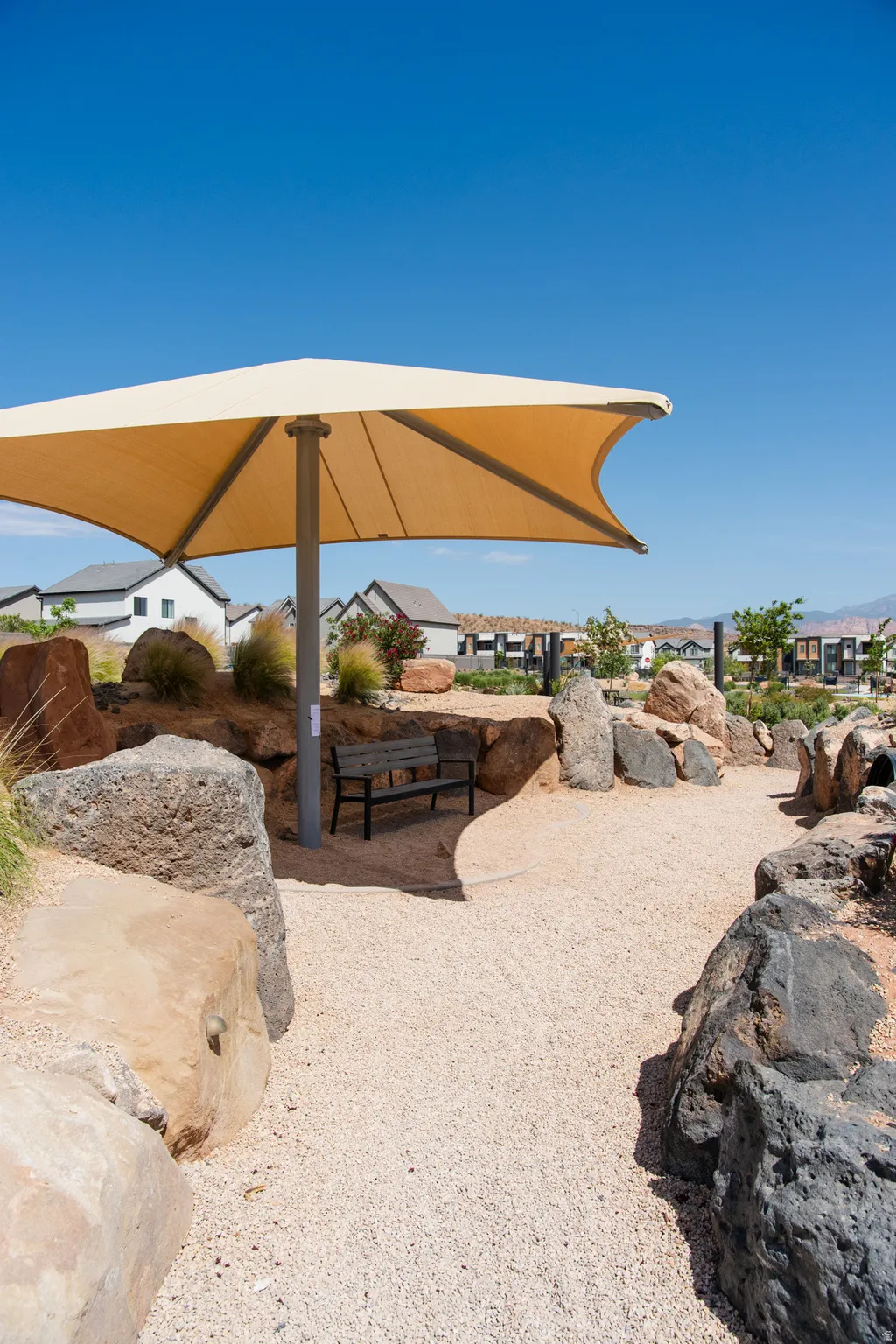 Shaded outdoor seating area featuring a beige sun sail, a dark wood-finish bench, and a gravel path