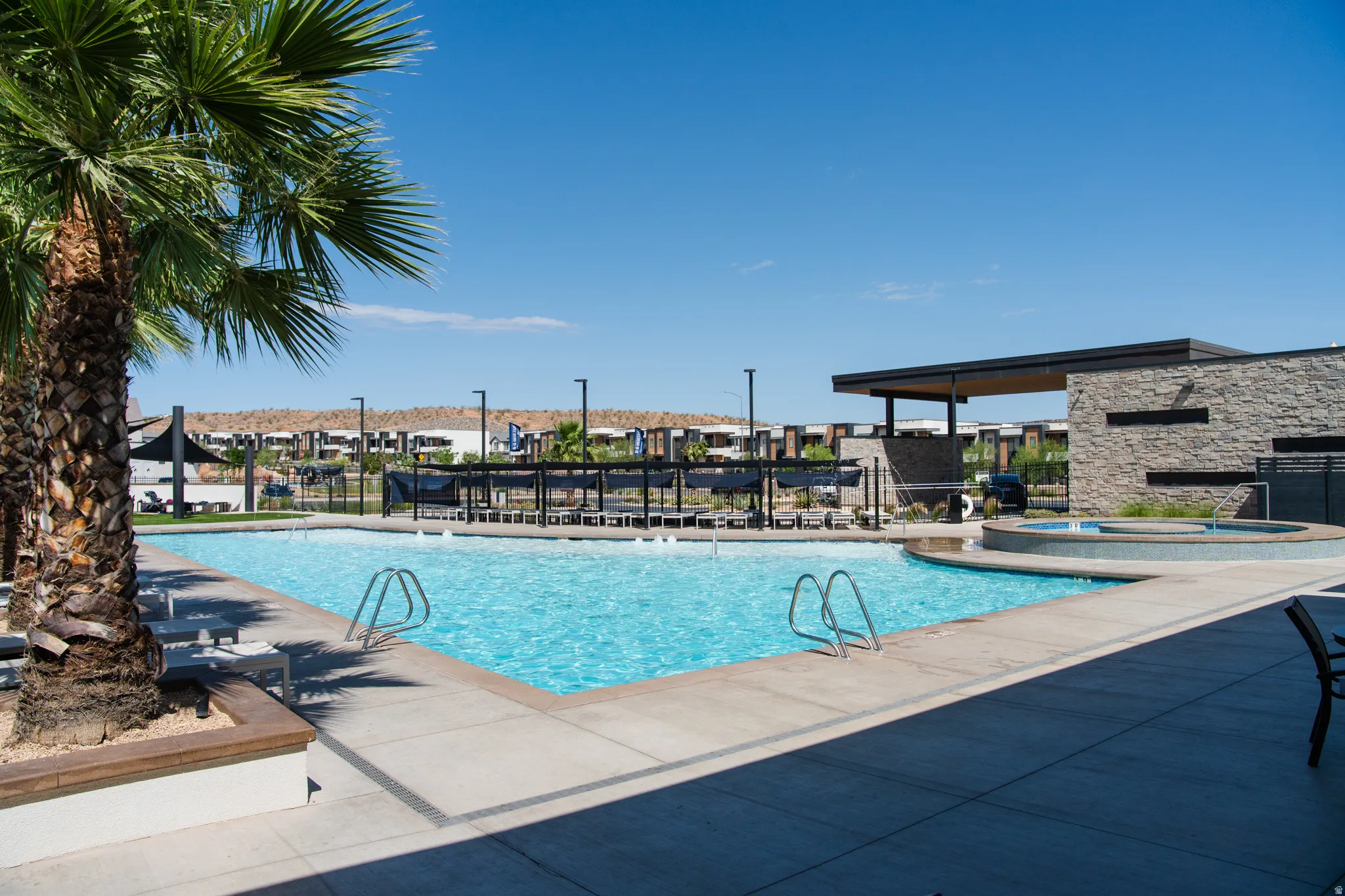 Resort-style resort pool and spa area featuring crystal-clear waters, built-in entry ladder, expansive sun deck with comfortable lounge seating, mature palm landscaping, modern shade structure with integrated pergola design, and adjacent stone architectur