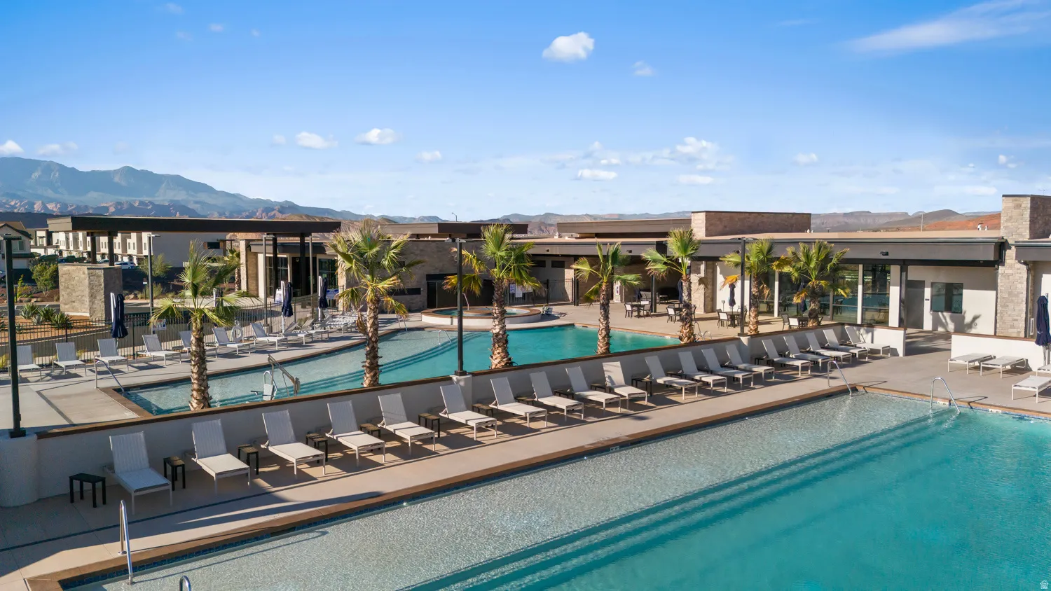 Resort-style resort pool and spa area featuring two sparkling swimming pools, an expansive sun deck lined with lounge seating, mature palm trees, and a modern clubhouse structure with covered pavilions