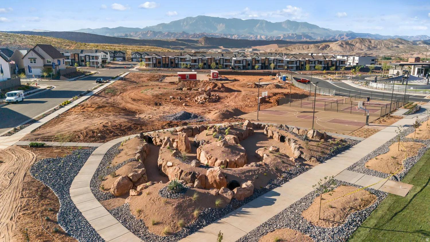 Community recreation area featuring a multi-court sports area, natural rock play structure, paved walking paths, landscaped green spaces, and mountain backdrop