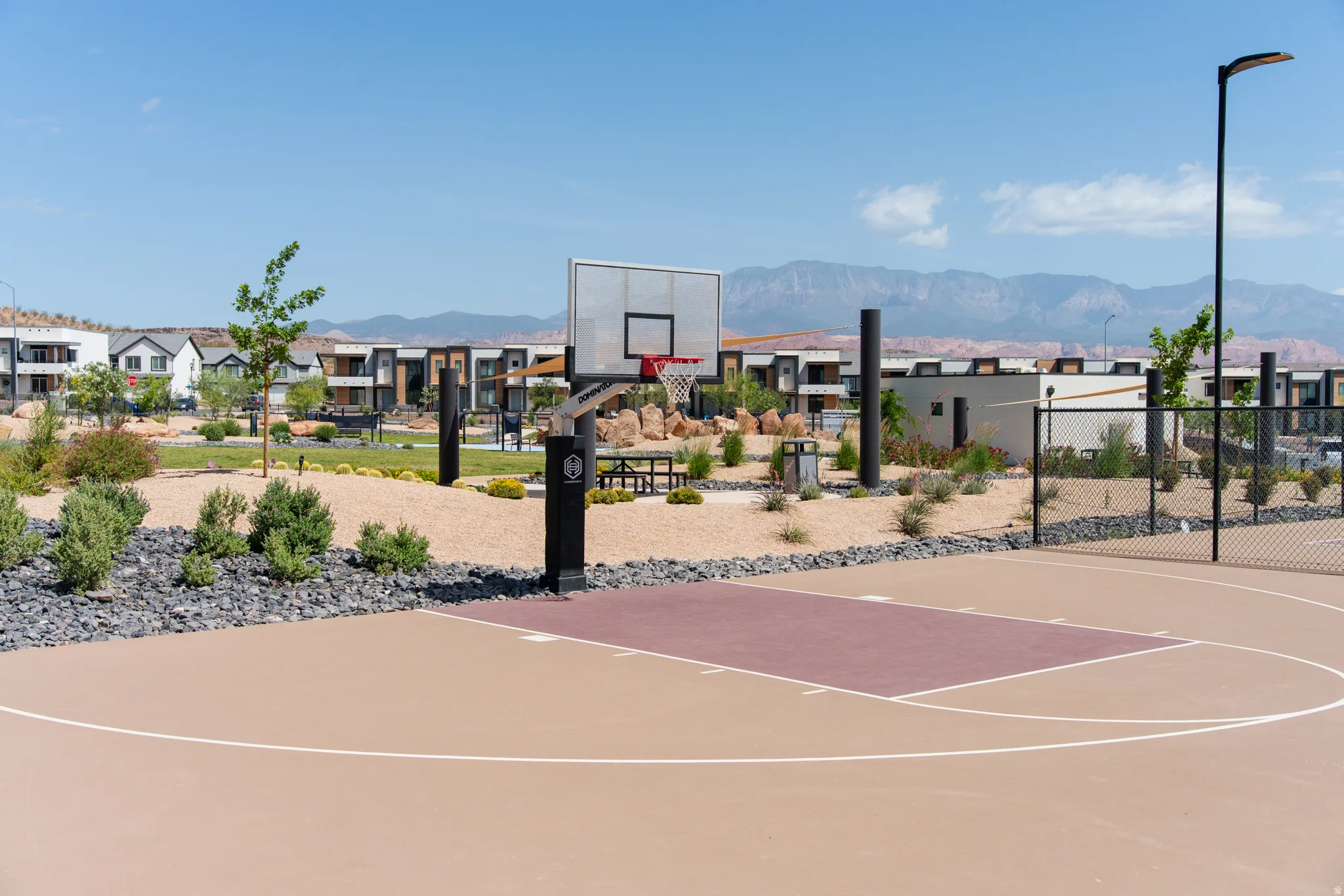 Outdoor basketball court with a tan and maroon playing surface, a black basketball hoop, and perimeter fencing