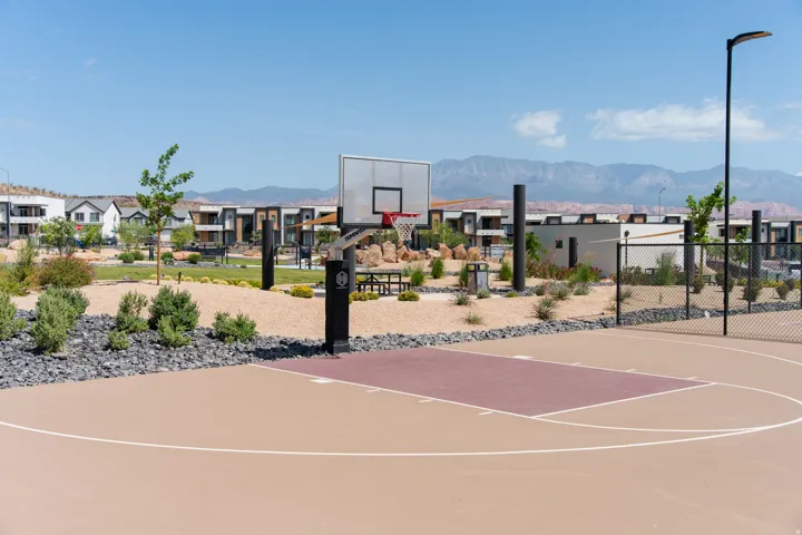 Outdoor basketball court with a tan and maroon playing surface, a black basketball hoop, and perimeter fencing