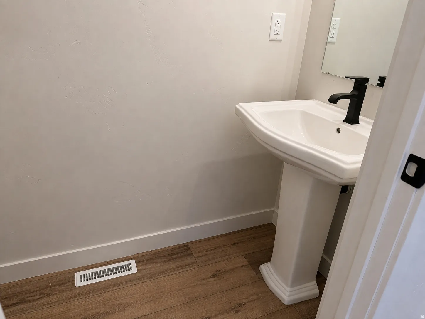 Pedestal sink with matte black faucet, wood-finish flooring, and crisp white baseboards