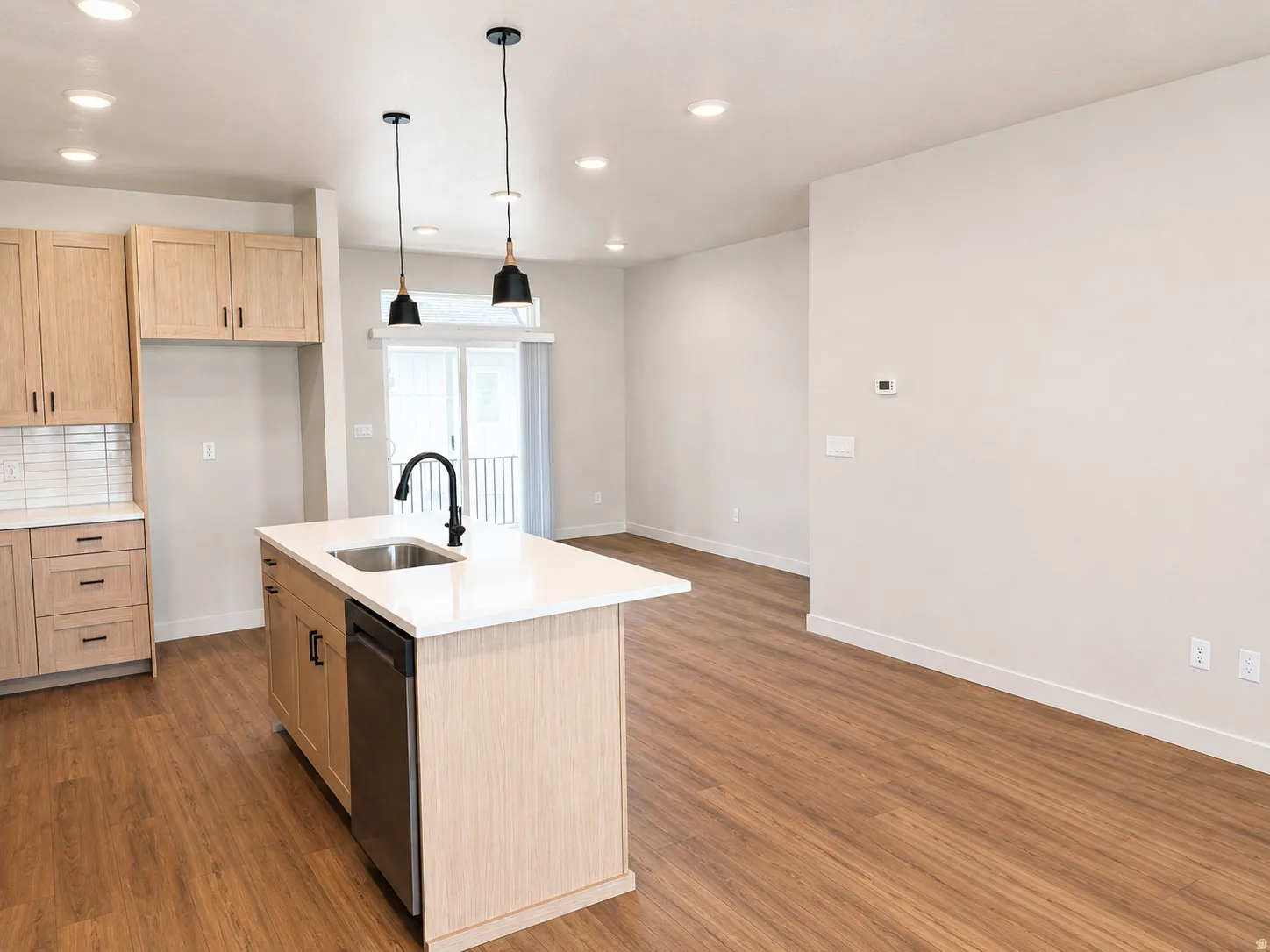 Modern kitchen featuring a substantial island with white quartz countertop and black matte faucet, light wood cabinetry throughout, stainless steel appliances, pendant lighting, recessed ceiling lights, and warm wood-finish flooring extending into the ope