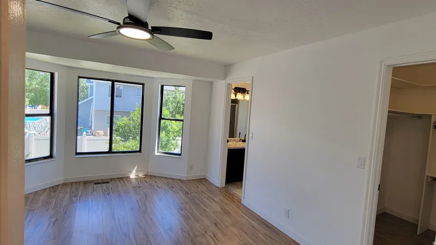Bay window with multiple large windows, wood-finish flooring, ceiling fan with integrated lighting, and an adjacent walk-in closet