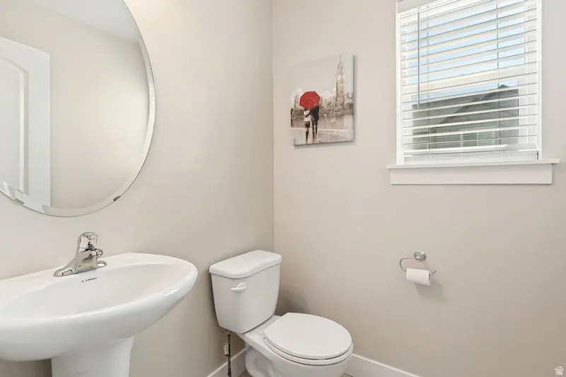 Bright powder room featuring a curved mirror with polished chrome fixtures, pedestal sink, white ceramic toilet, horizontal blinds, and neutral wall finishes