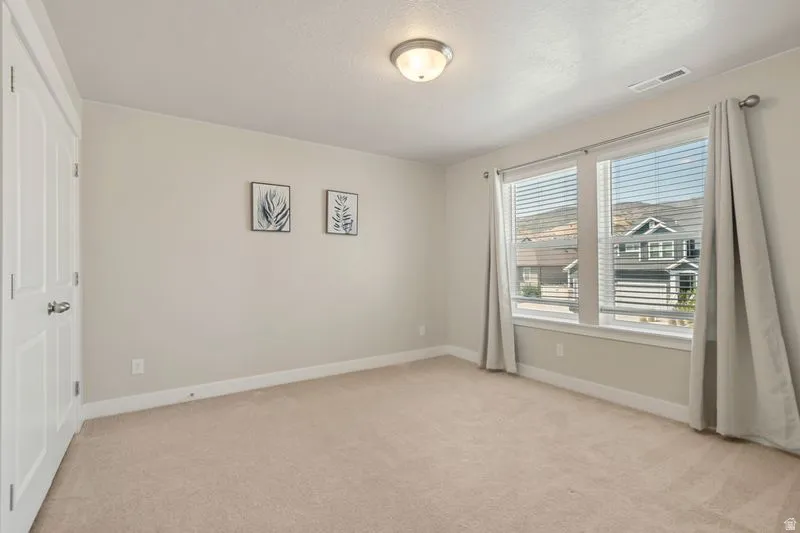 Spacious bedroom featuring neutral carpeting, abundant natural light from dual large windows with custom drapery, ceiling-mounted lighting fixture, and direct access to a closet