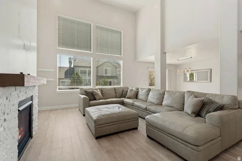 Spacious living area featuring high ceilings, large windows with blinds, a fireplace with a stone surround and wood mantel, and light wood-finish flooring