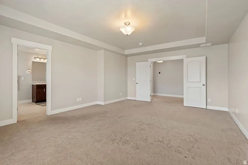 Spacious primary suite featuring neutral carpeting, multiple closet entries with white six-panel doors, recessed ceiling detail with crown molding, and abundant natural light from ceiling fixture