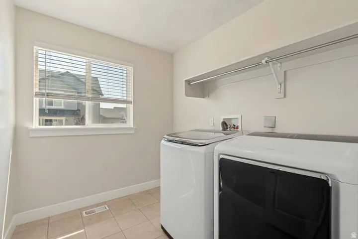 Dedicated laundry room featuring side-by-side washer and dryer appliances, tile flooring, ample counter space with stainless steel rod, and natural light from a double-hung window with plantation shutters