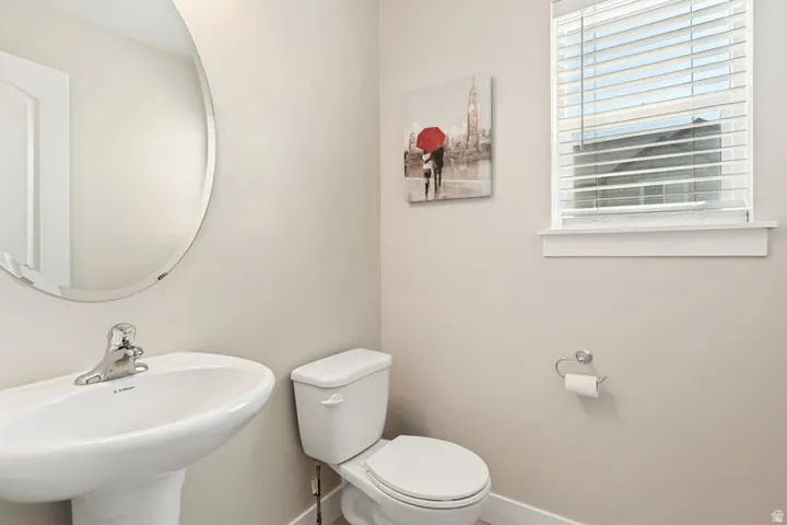 Bright powder room featuring a curved mirror with polished chrome fixtures, pedestal sink, white ceramic toilet, horizontal blinds, and neutral wall finishes