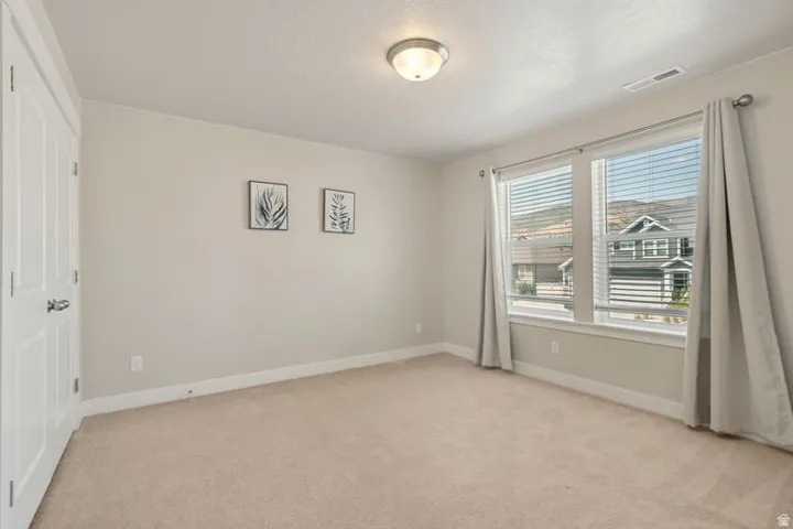Spacious bedroom featuring neutral carpeting, abundant natural light from dual large windows with custom drapery, ceiling-mounted lighting fixture, and direct access to a closet
