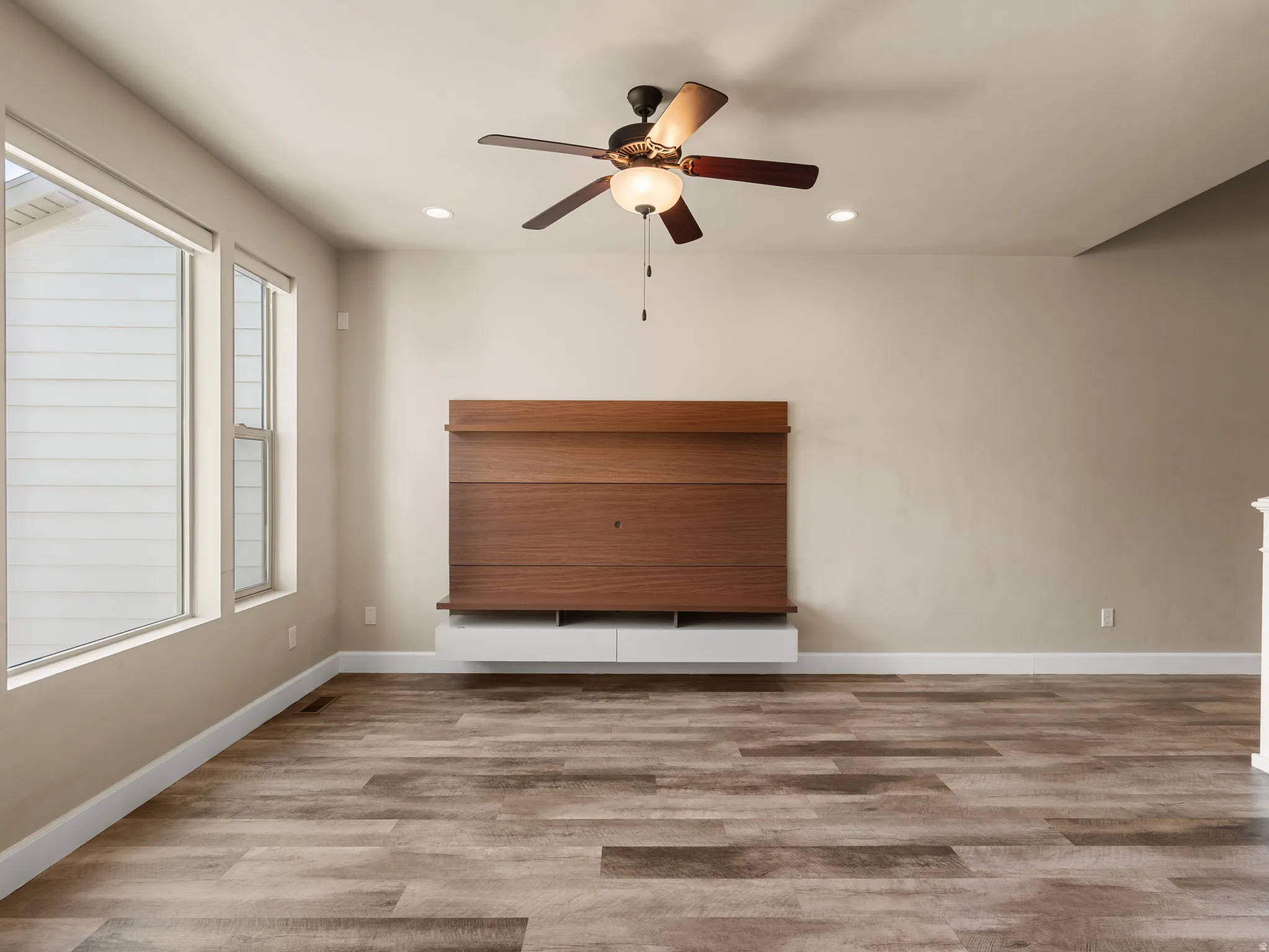 Spacious room featuring wood-finish flooring, a built-in media console with a wood panel accent, and a ceiling fan with integrated lighting