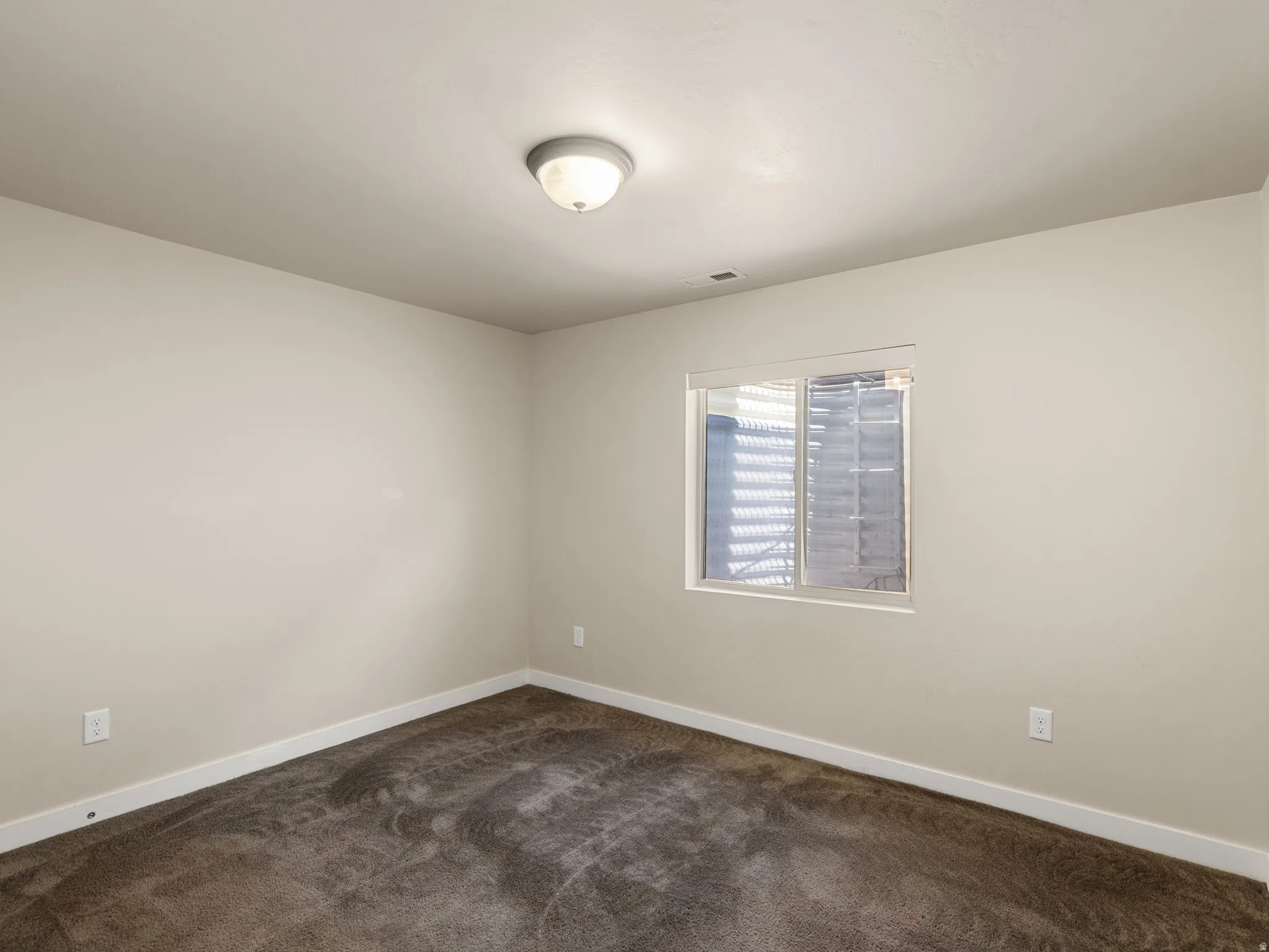 Spacious bedroom featuring neutral wall finishes, carpeted flooring, dual wall-mounted outlets, recessed ceiling lighting, and a window with integrated blinds providing natural light