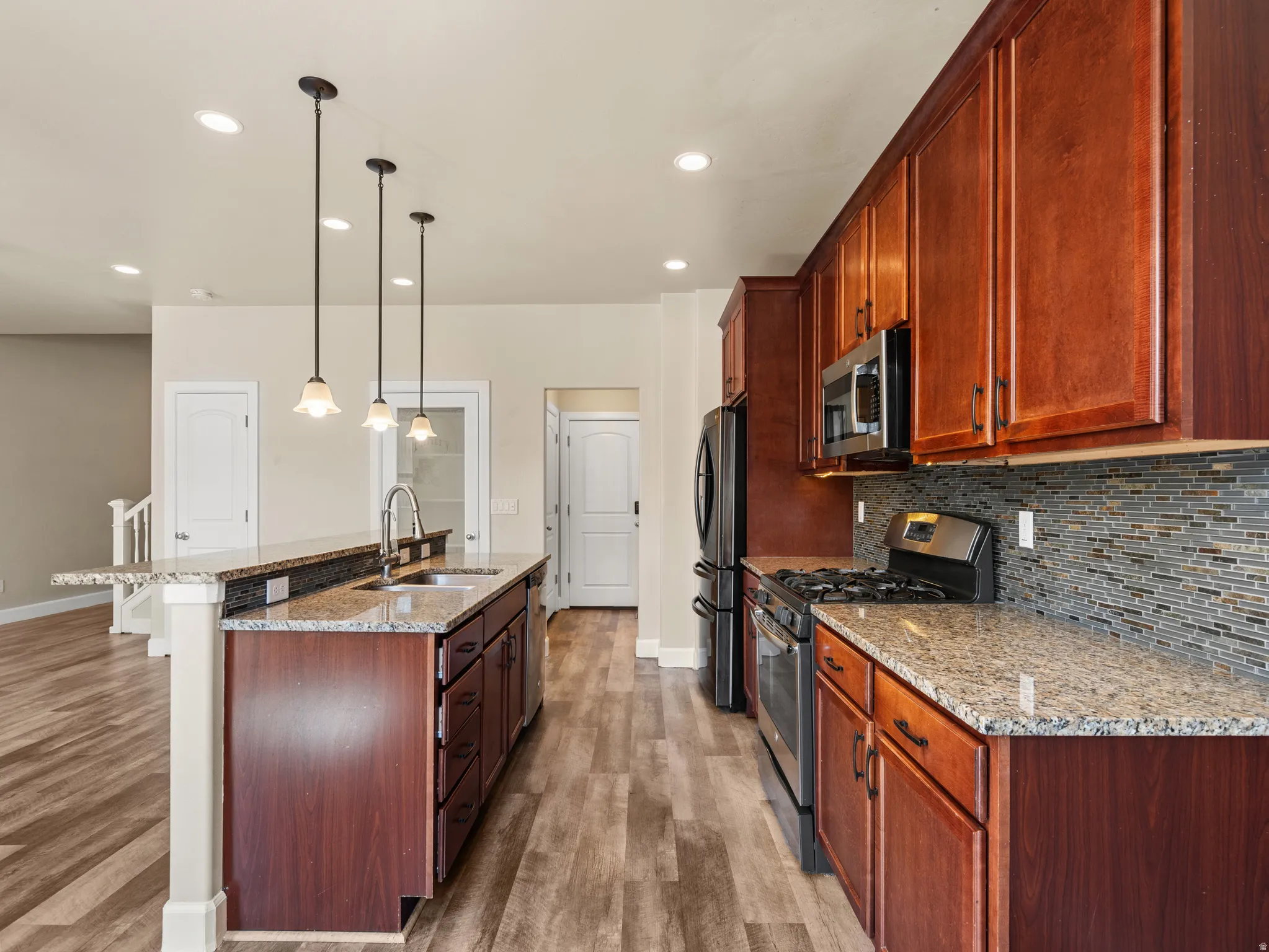 Kitchen featuring an island with a built-in sink, pendant lighting, wood-finish flooring, granite countertops, and stainless steel appliances