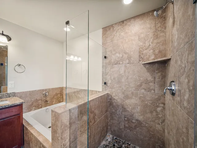 Bathroom featuring a tiled walk-in shower with a built-in shelf, a separate soaking tub, and a vanity with a granite-style countertop