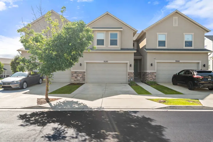 Stucco exterior featuring stacked stone accents, two single-car garages with concrete driveways, and a mature tree providing natural shade