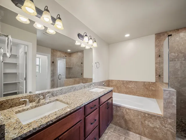 Spa-inspired primary bathroom featuring dual vanities with granite countertops, warm wood cabinetry, and brushed nickel hardware