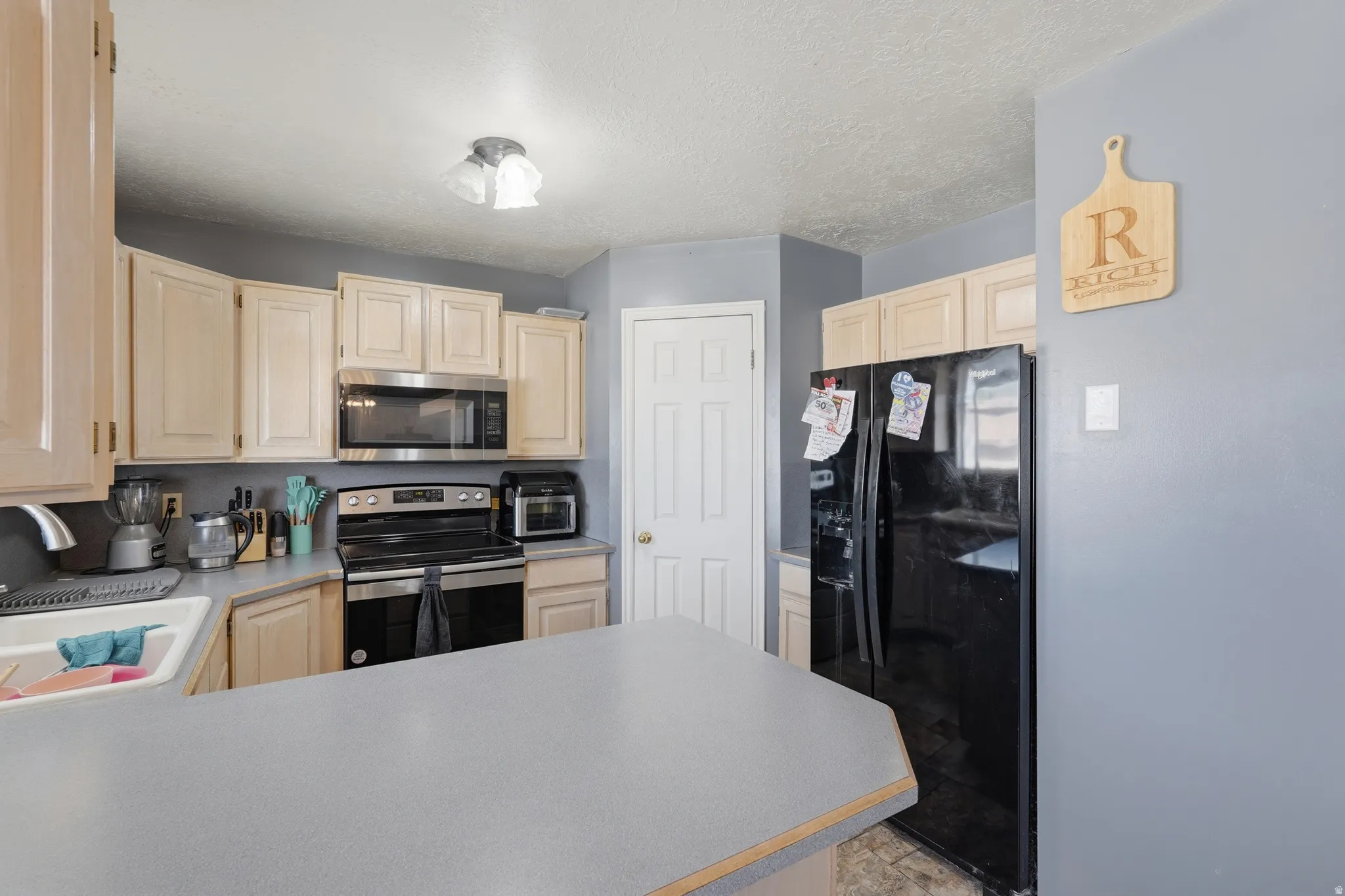 Bright kitchen featuring light wood cabinetry, stainless steel appliances including a black refrigerator and range, white quartz countertops with breakfast bar seating, and neutral wall finishes