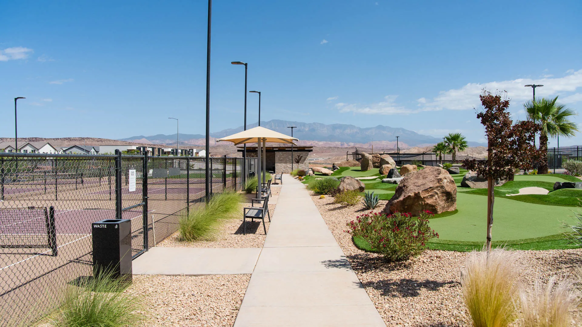 Community park area featuring a concrete walking path, manicured lawn with native landscaping, mature palm trees, and modern street lighting