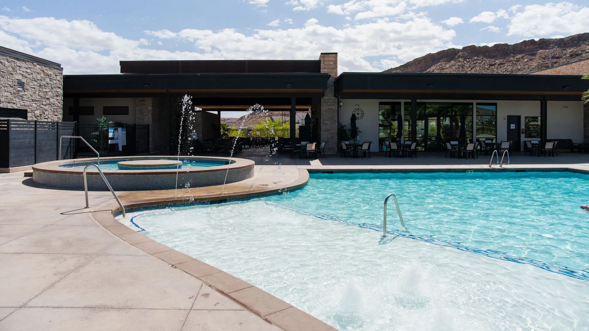 Outdoor swimming pool with integrated spa and water features, expansive pool deck, and a contemporary clubhouse building featuring large glass panels and a covered patio area with seating