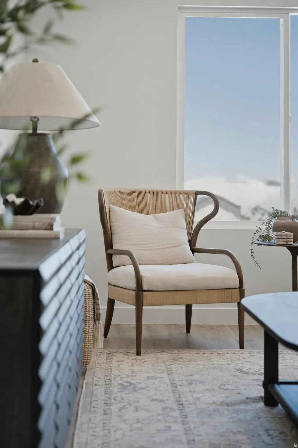 Bright corner room featuring floor-to-ceiling windows with expansive natural light, wood-frame wingback accent chair with cream upholstery, neutral area rug, and contemporary side table