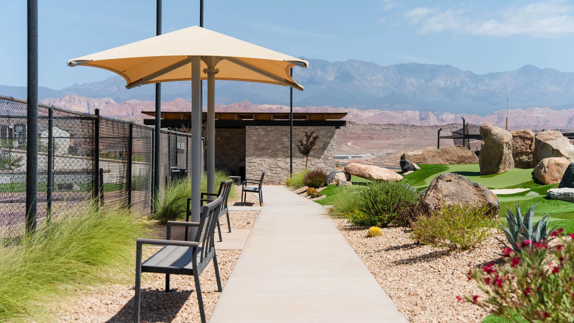 Outdoor entertaining space featuring a tan shade structure with integrated seating area, concrete walkway, and professionally landscaped grounds with native plantings, decorative rock features, and manicured lawn