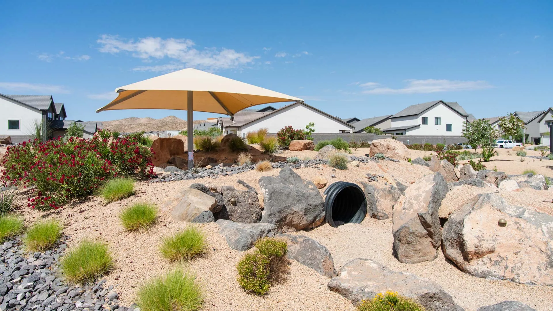 Desert-style landscaping featuring a shade structure, natural boulders, red flowering shrubs, and low-maintenance plantings