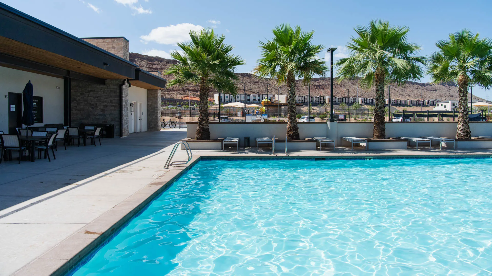 Sparkling outdoor swimming pool featuring a concrete deck, multiple palm trees, and a stone-accented building with an open-air patio
