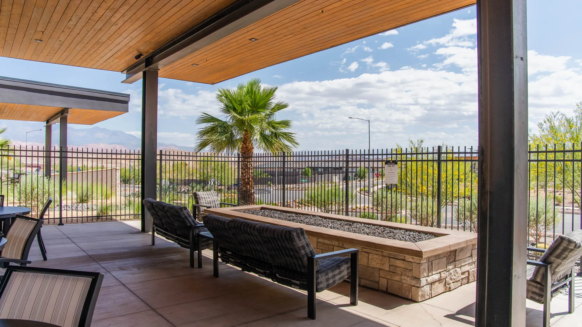 Expansive covered patio with wood-finish ceiling and steel support columns, featuring a built-in brick bar with seating, lounge chairs, and ornamental metal railing
