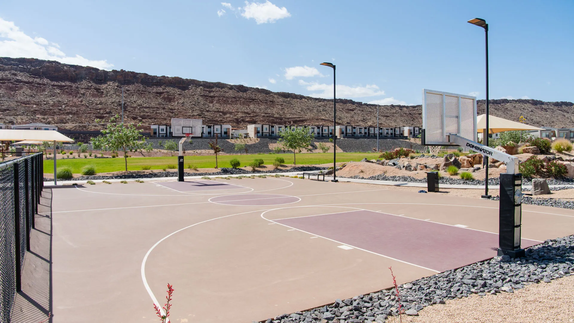 Outdoor multi-court surface featuring two basketball hoops with backboards, surrounding landscape with green space, and overhead lighting fixtures