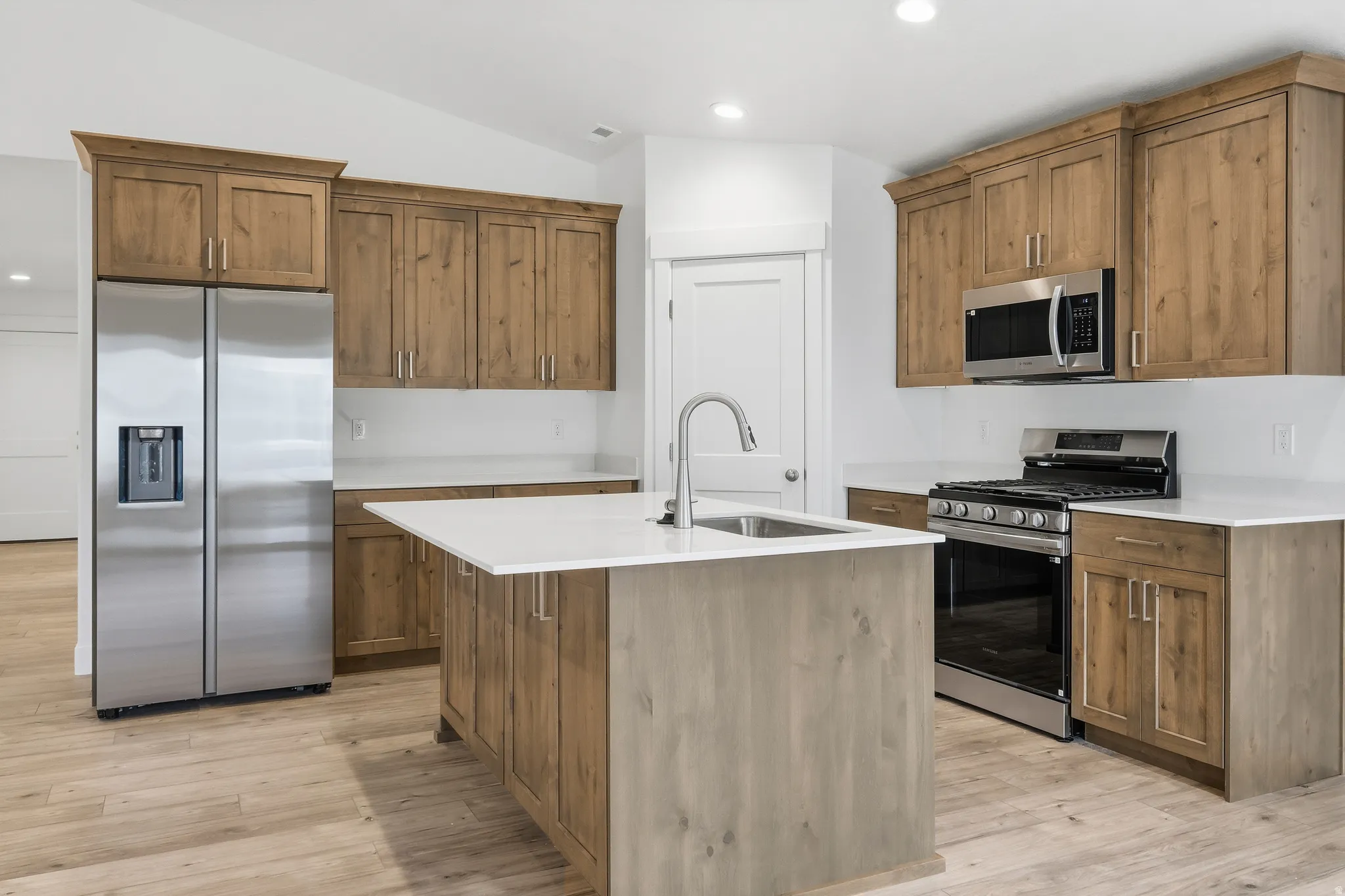 Modern kitchen featuring light wood-finish cabinetry, stainless steel appliances, a center island with a sink, white countertops, and wood-finish flooring
