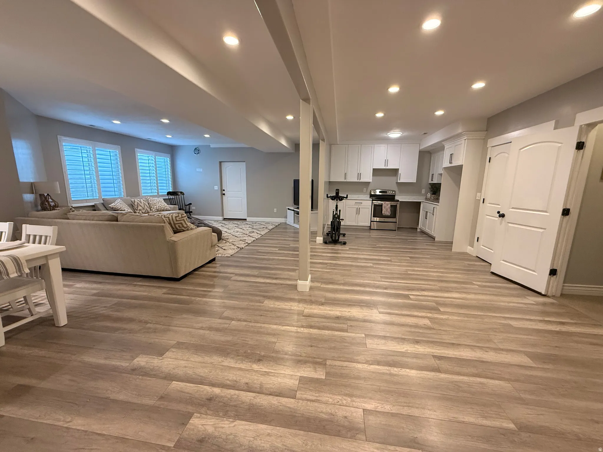 Recessed lighting illuminates the open-concept space featuring wood-finish flooring throughout, a full kitchen with white cabinetry, and two paneled doors