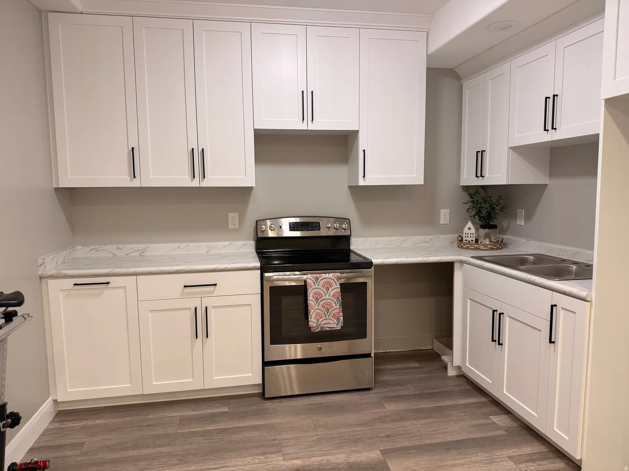 White shaker cabinetry with matte black hardware, stainless steel electric range, double basin stainless steel sink, wood-finish flooring, and light-toned countertops