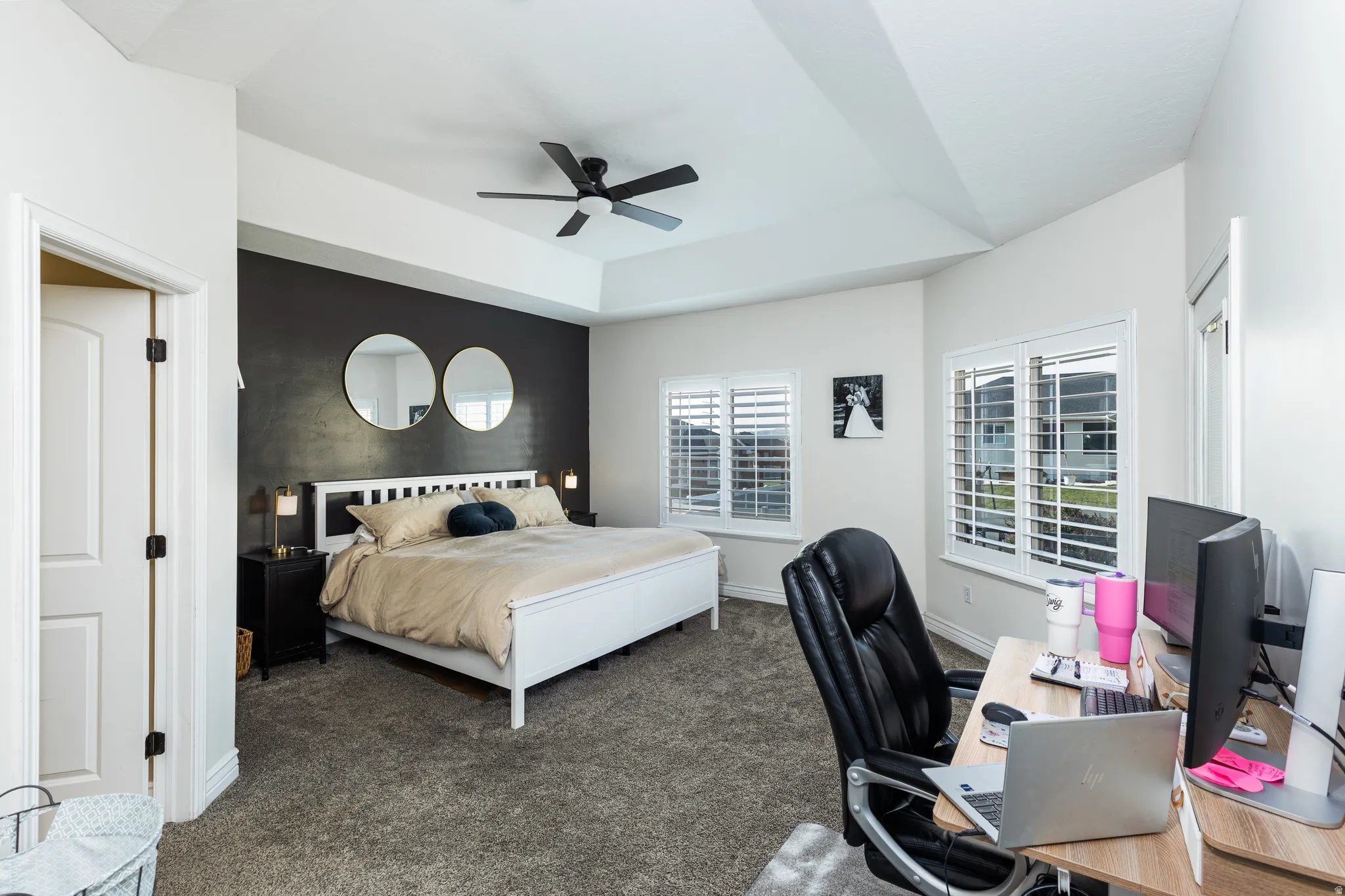 Vaulted ceiling with a black ceiling fan, dark accent wall, two large windows with plantation shutters, light-toned carpeting,