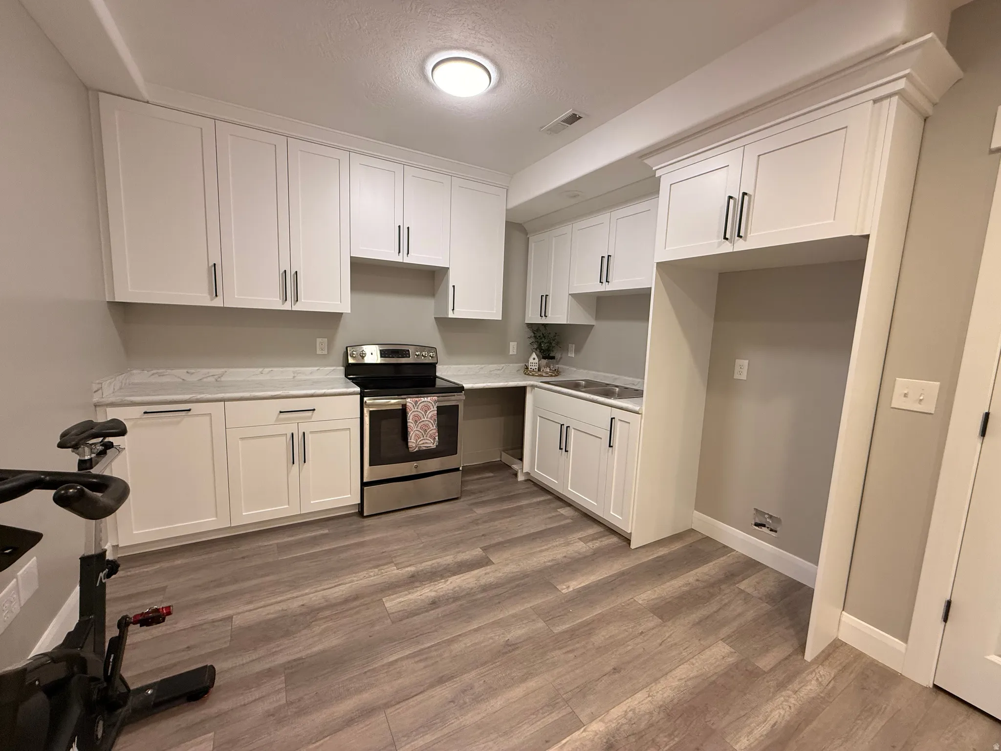 White shaker-style cabinetry with black hardware, stainless steel electric range, undermount stainless steel double sink, light-toned countertops, and wood-finish flooring