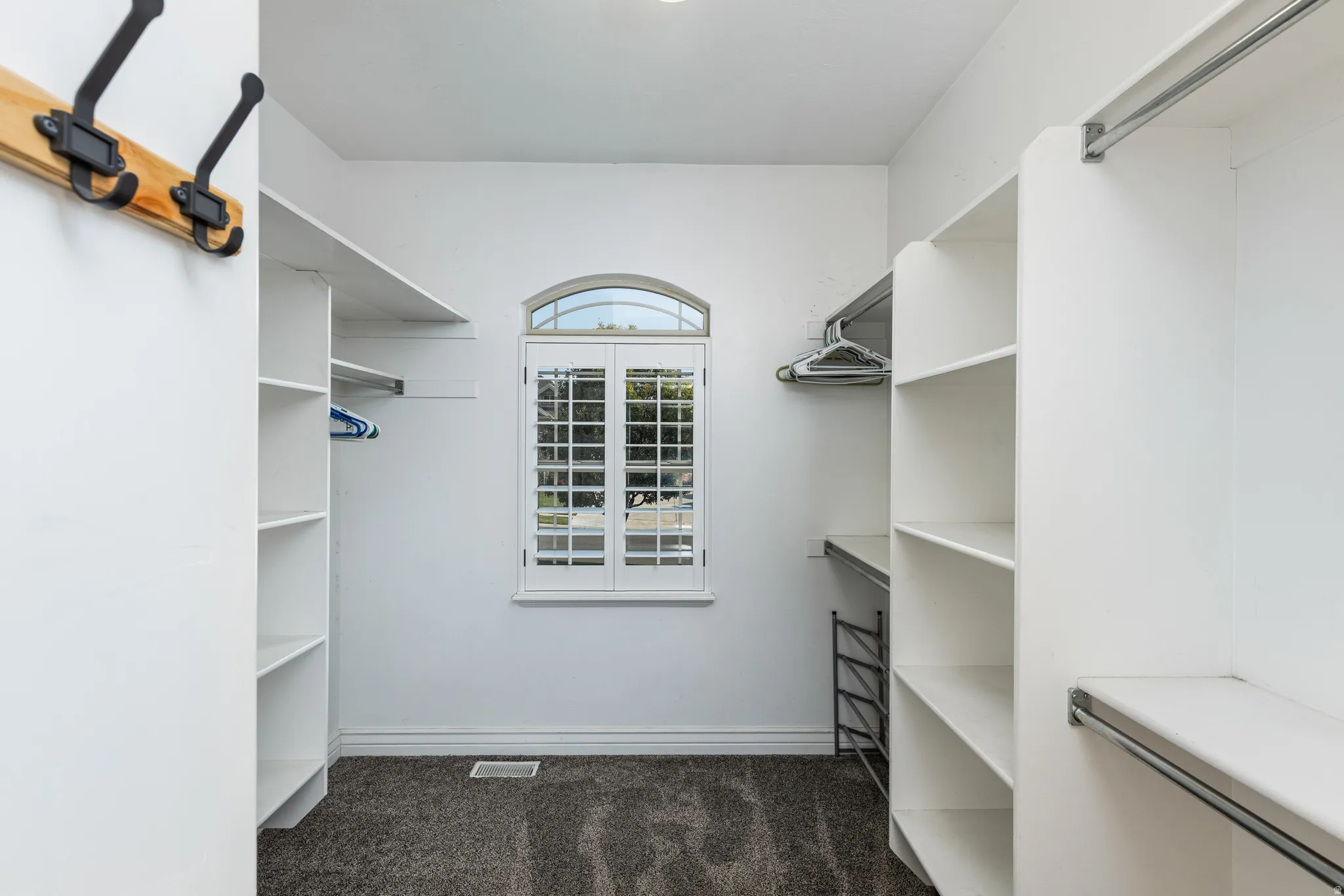 Walk-in closet featuring an arched window with white plantation shutters, built-in shelving, hanging rods, and dark gray carpet