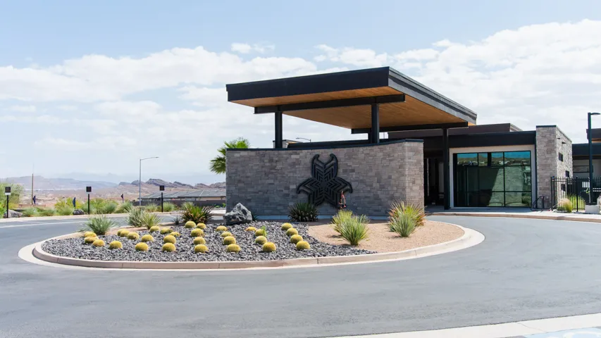 Modern community entrance featuring a stone-clad gatehouse with a cantilevered roof, desert landscaping, and a central roundabout