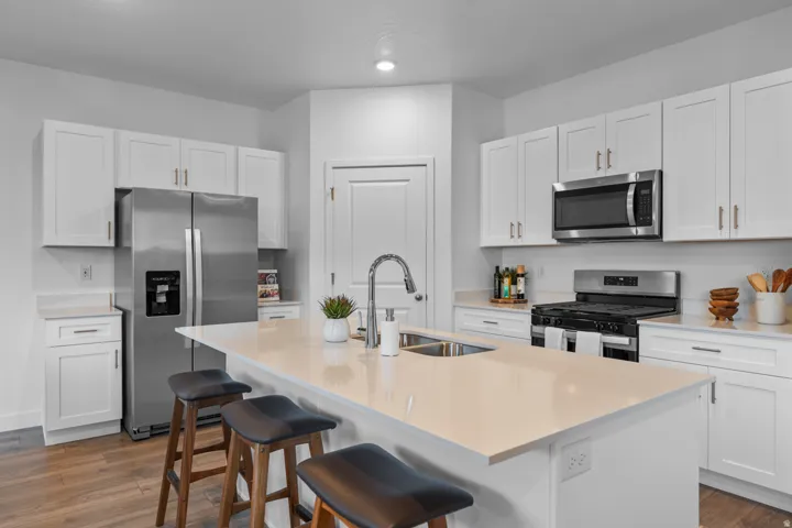 Kitchen featuring a central island with integrated sink, stainless steel appliances, white cabinetry with brass-finish hardware, light-toned countertops, and wood-finish flooring