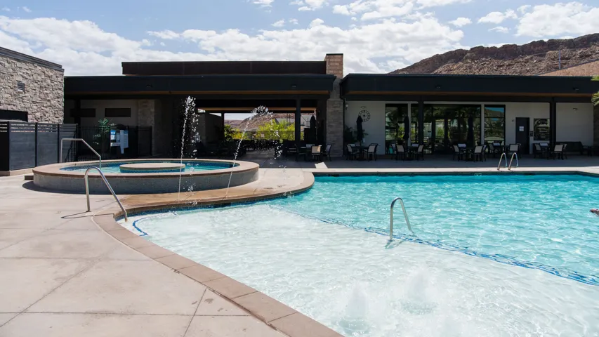 Outdoor swimming pool with integrated spa and water features, expansive pool deck, and a contemporary clubhouse building featuring large glass panels and a covered patio area with seating