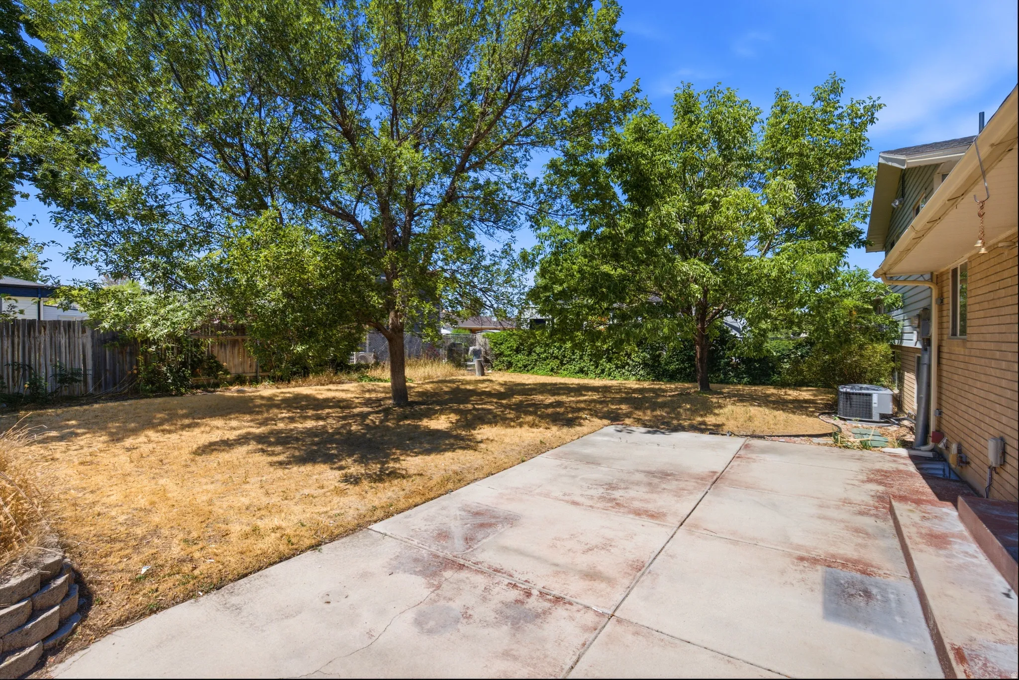Expansive backyard featuring a concrete patio, mature trees, and a retaining wall