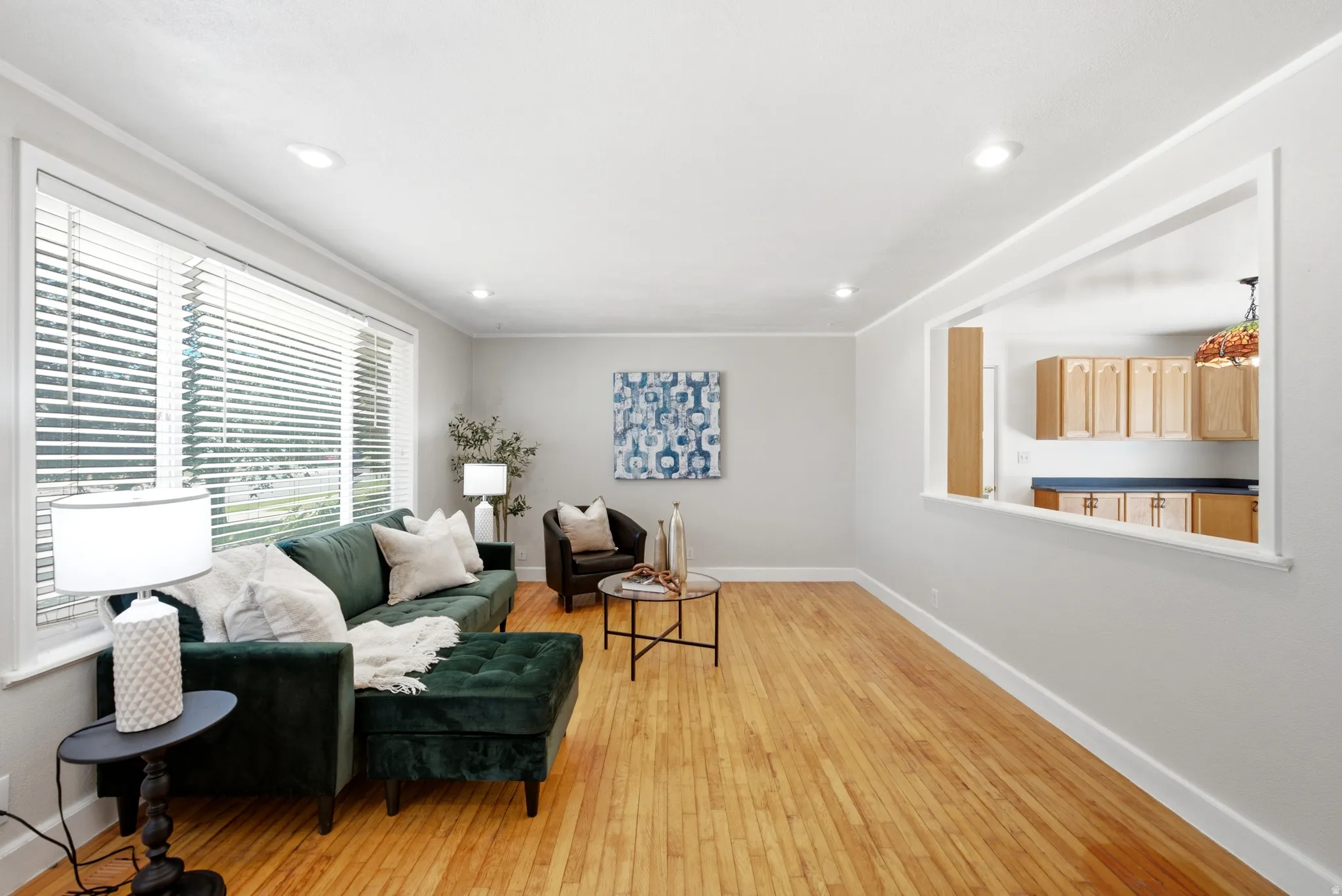 Light wood-finish flooring throughout the room, recessed ceiling lighting, pass-through opening to the kitchen, and a large window with white blinds