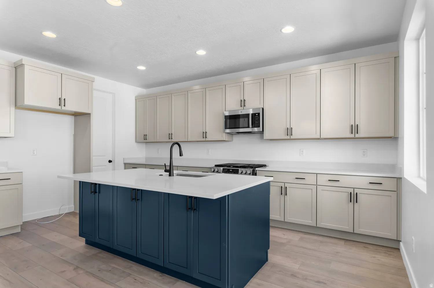 Kitchen featuring a blue island with integrated sink, stainless steel microwave, light-toned cabinetry, light-toned countertops, and wood-finish flooring