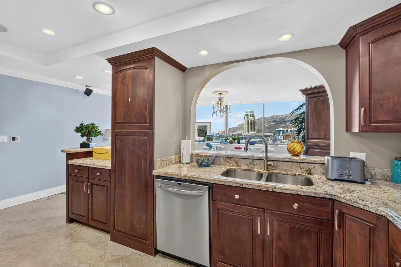 Kitchen featuring an arched pass-through window with city and mountain scenery, wood-finish cabinetry, light-toned granite countertops, a double basin stainless steel sink, and stainless steel dishwasher