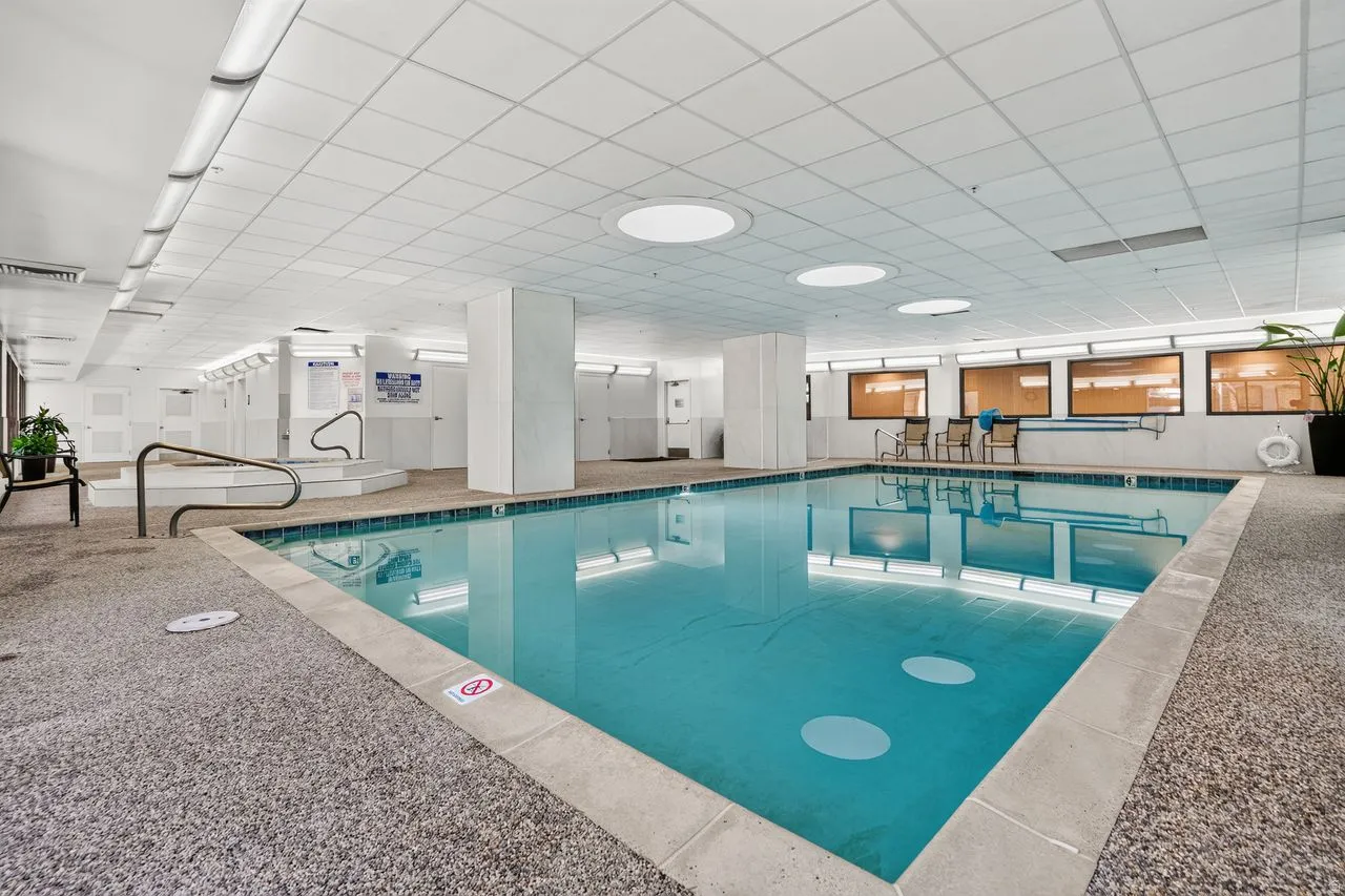 Indoor swimming pool featuring light blue tile, surrounding aggregate flooring, a raised spa with handrails, recessed ceiling lighting, and a drop ceiling